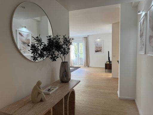 Ein heller Flur mit rundem Spiegel, Holzregal und Blumen, linker Zugang zu einem Raum.