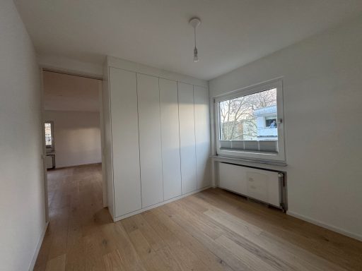 Helle, leere Wohnung mit Holzfußboden und Fenster, dahinter ein geschlossener Raum.