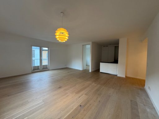 Heller, leerer Raum mit Holzboden und großen Fenstern, orangefarbene Pendelleuchte.